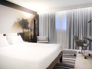 A hotel room with a large bed, a desk with a chair, a view of the outdoors through sheer curtains, and a painting on the wall.