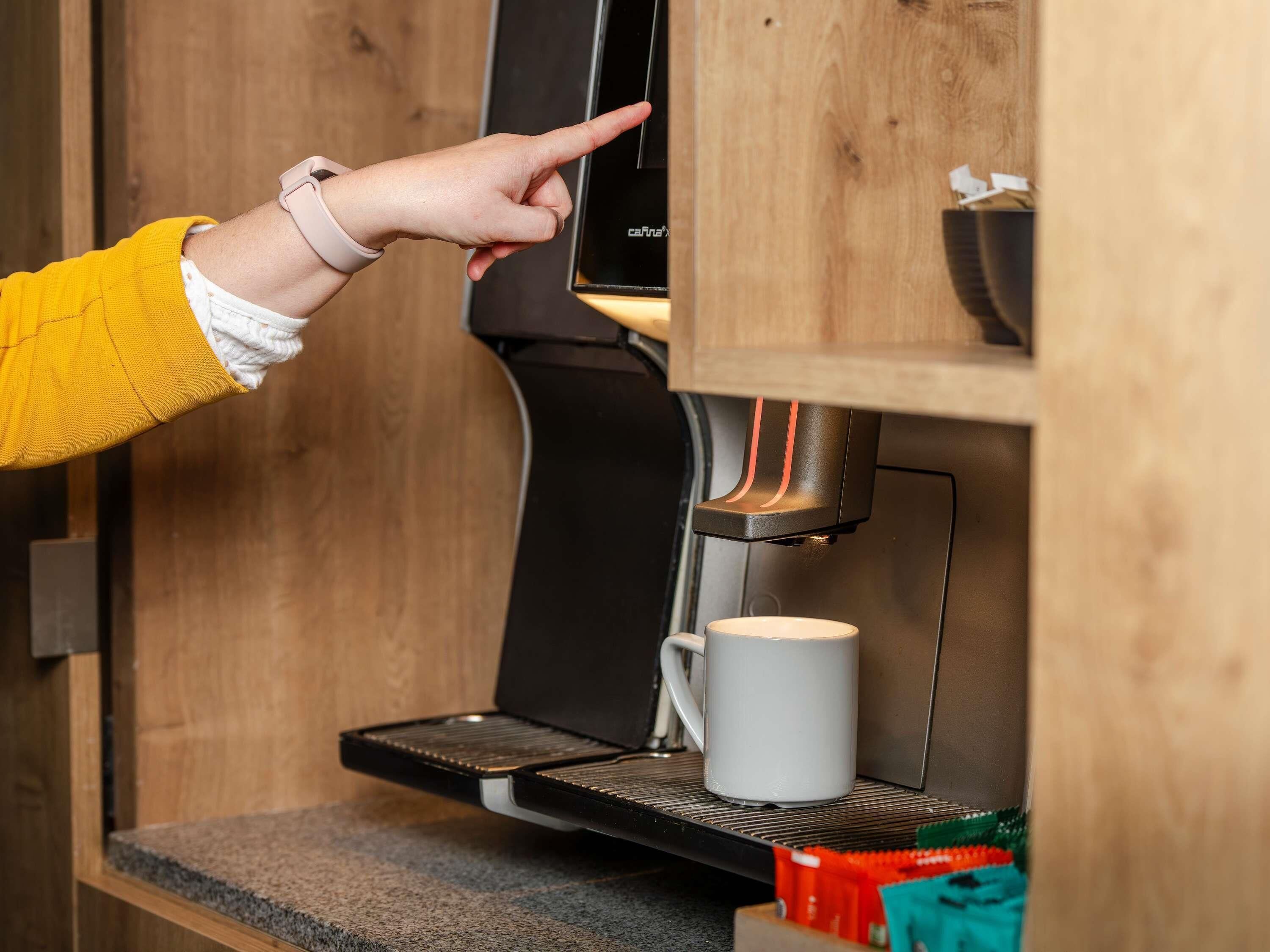 A coffee machine with a digital display, a coffee cup, and a person pointing at the display.