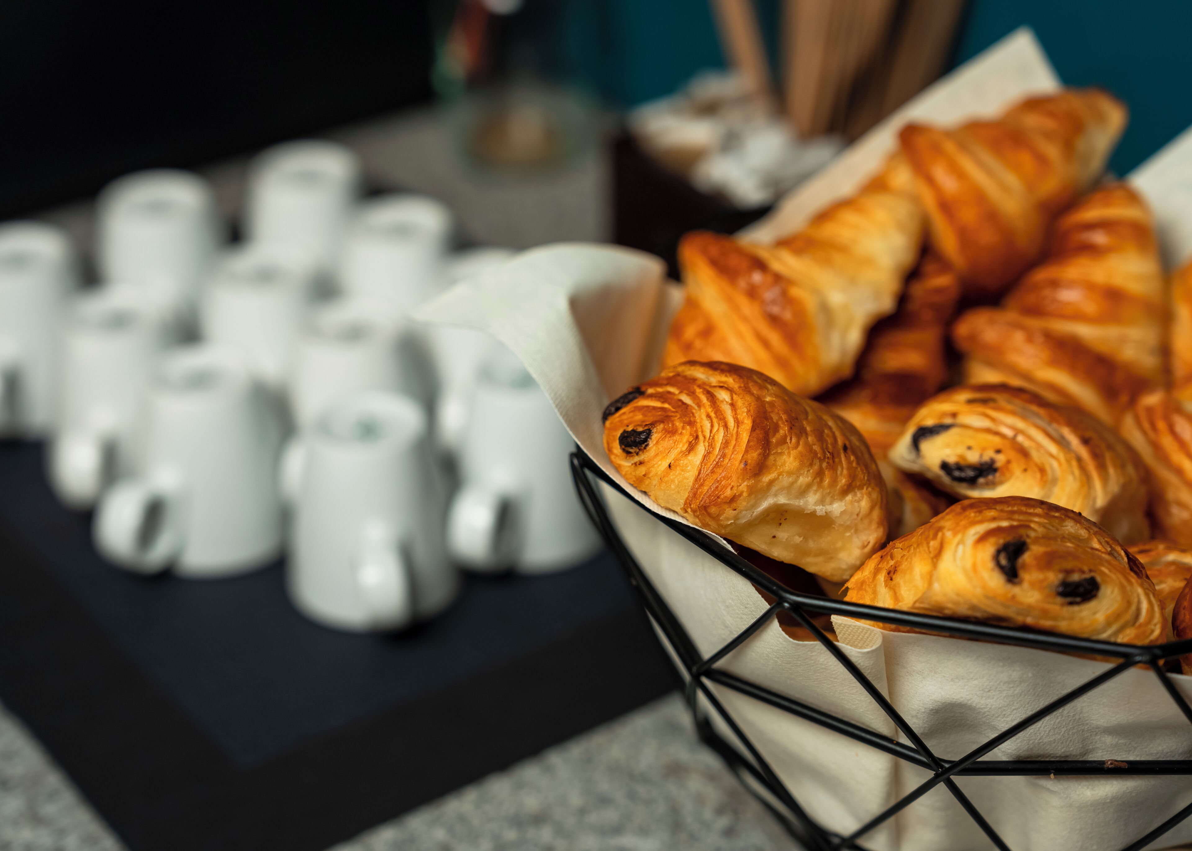 A basket of croissants and a row of white ceramic cups on a table.