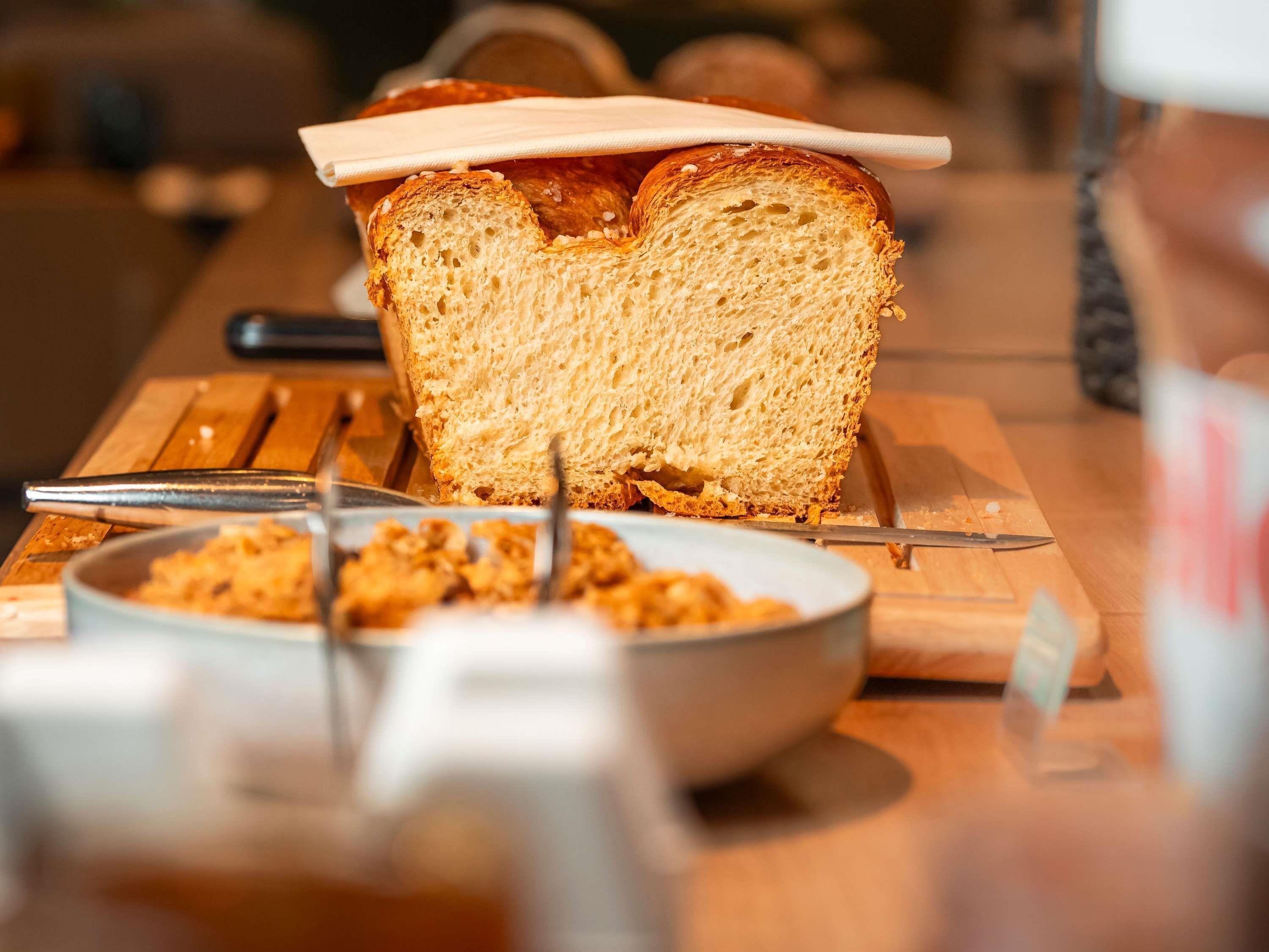 A loaf of bread on a wooden cutting board with a knife and a bowl of cereal.