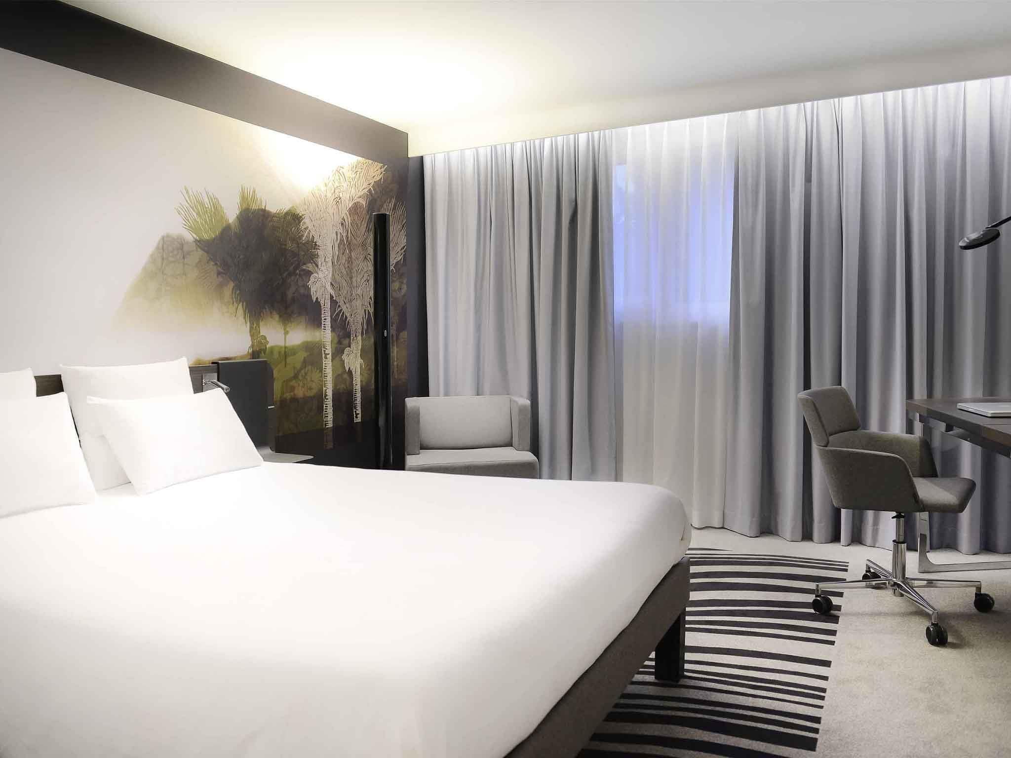 A hotel room with a large bed, two chairs, a desk, and a view of the outdoors through sheer curtains.