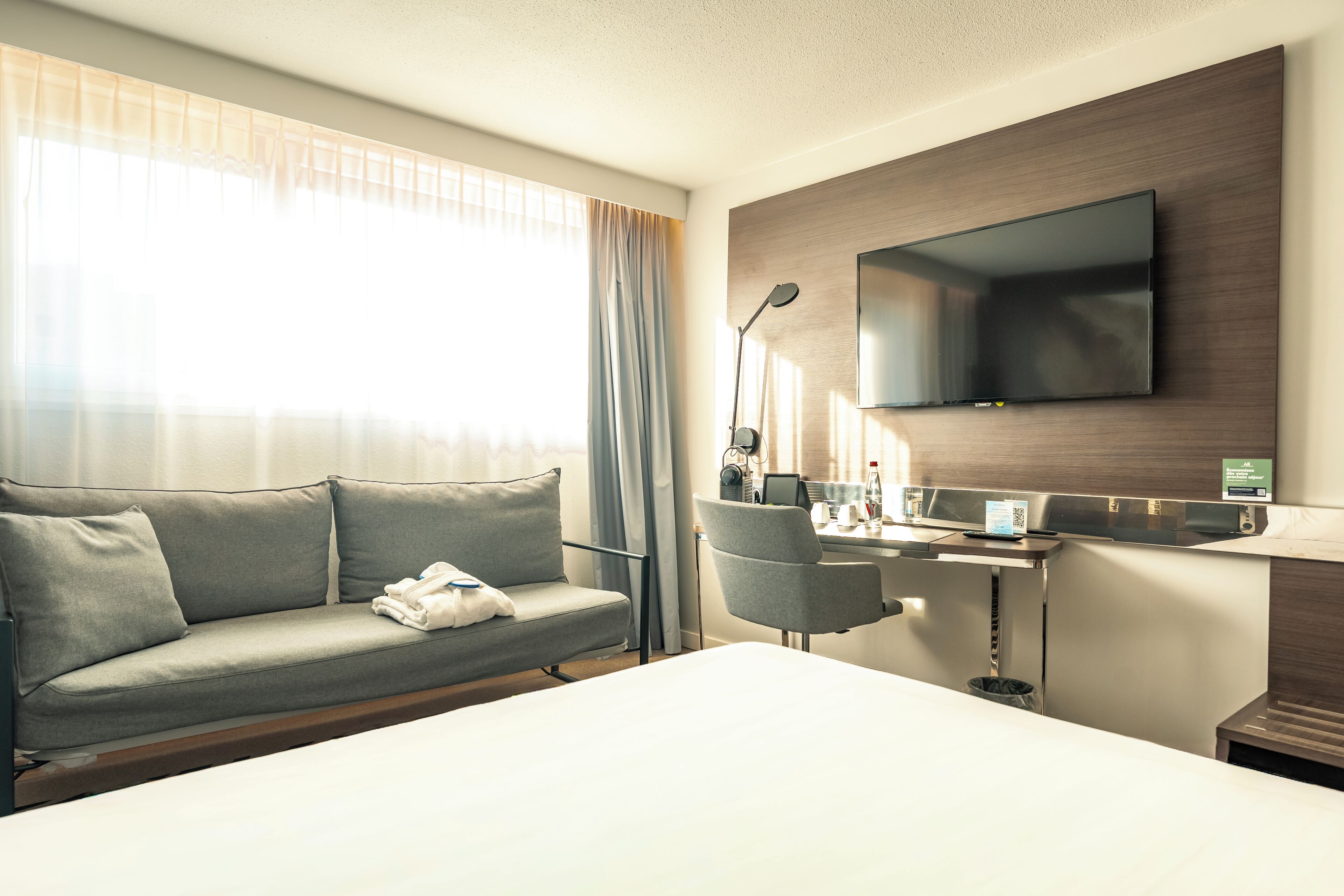 A modern hotel room with a large bed, a grey sofa, a desk with a flat-screen TV, and a window with sheer curtains.