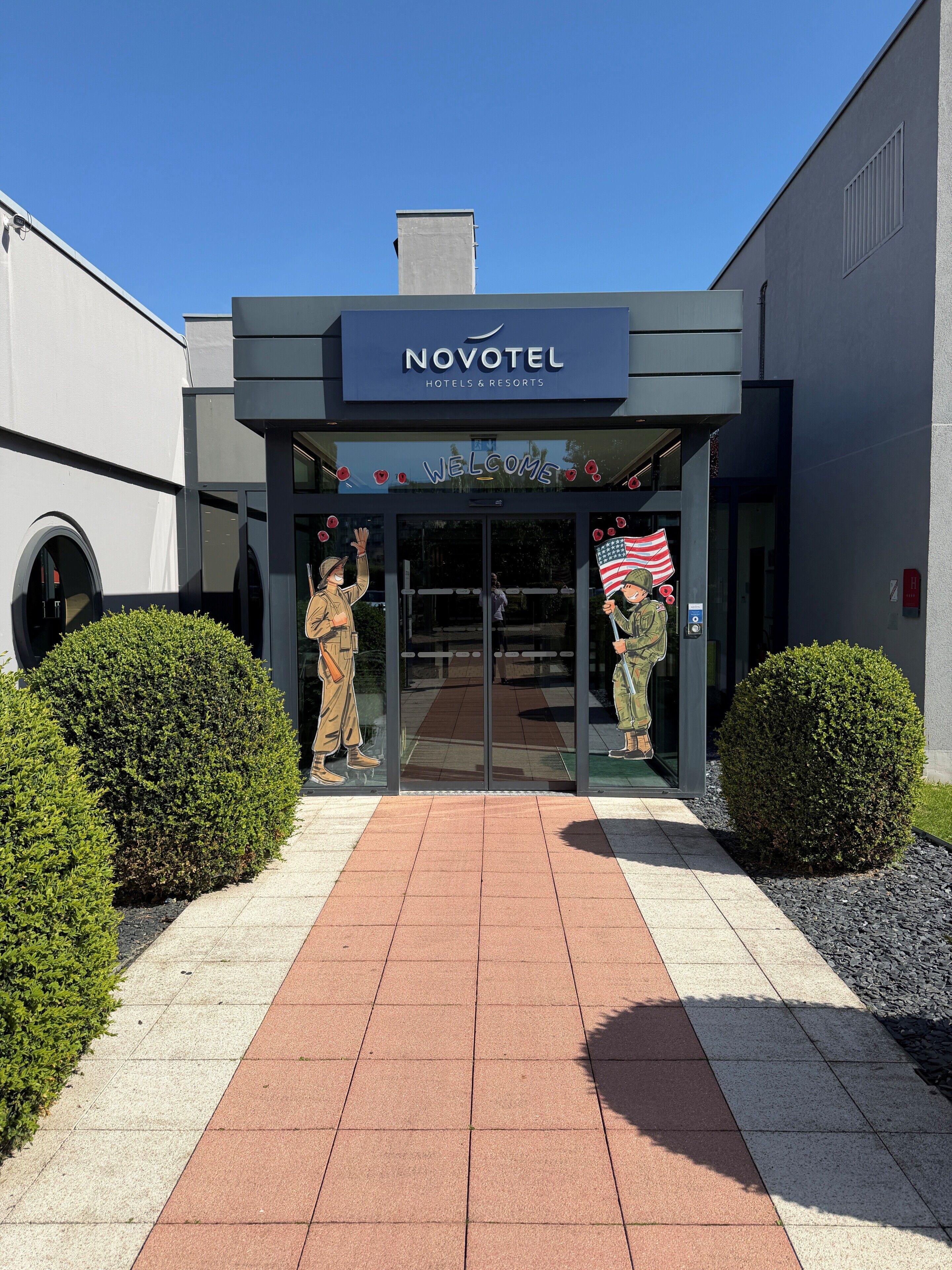 A modern hotel entrance with a sign reading 'NOVOTEL Hotels & Resorts,' a welcome sign, and two military-themed statues.