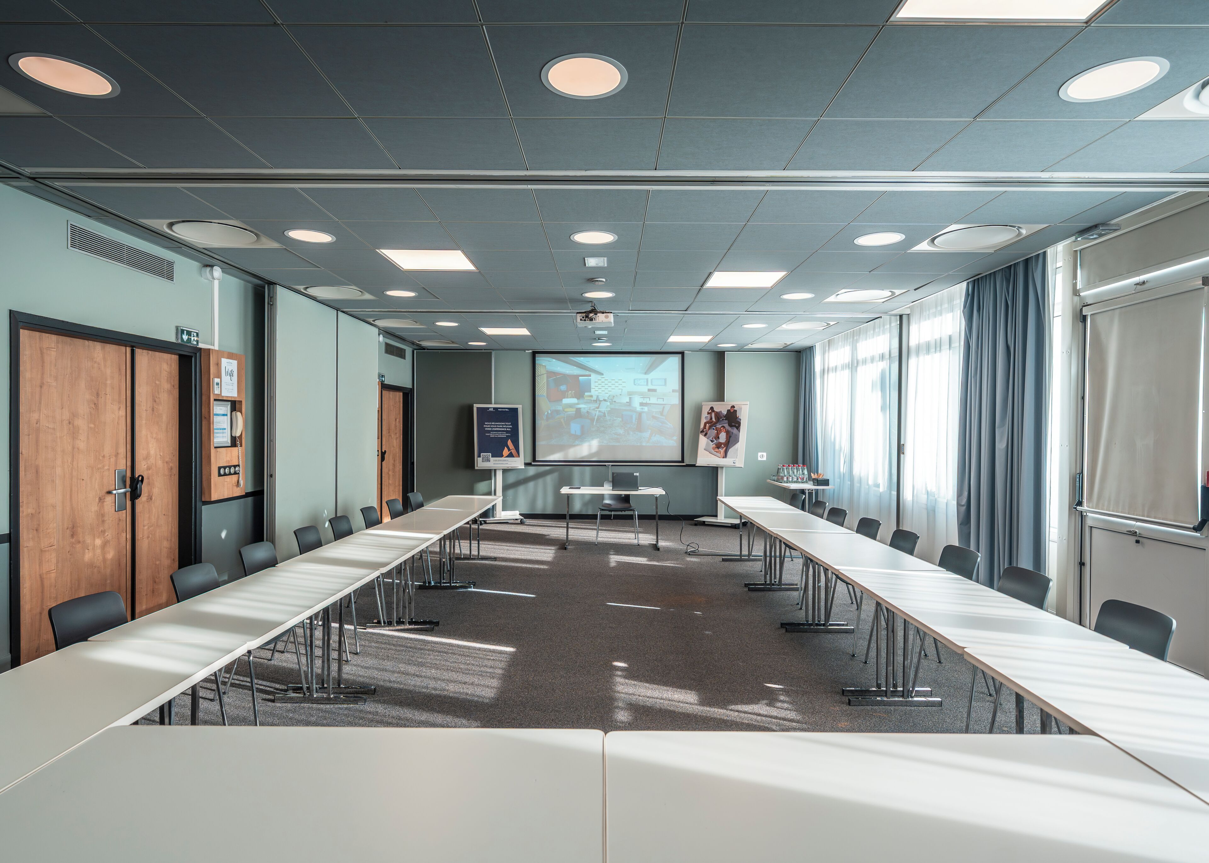 Un salón de conferencias con una larga mesa, sillas, una pantalla de proyector y una pared para presentaciones.