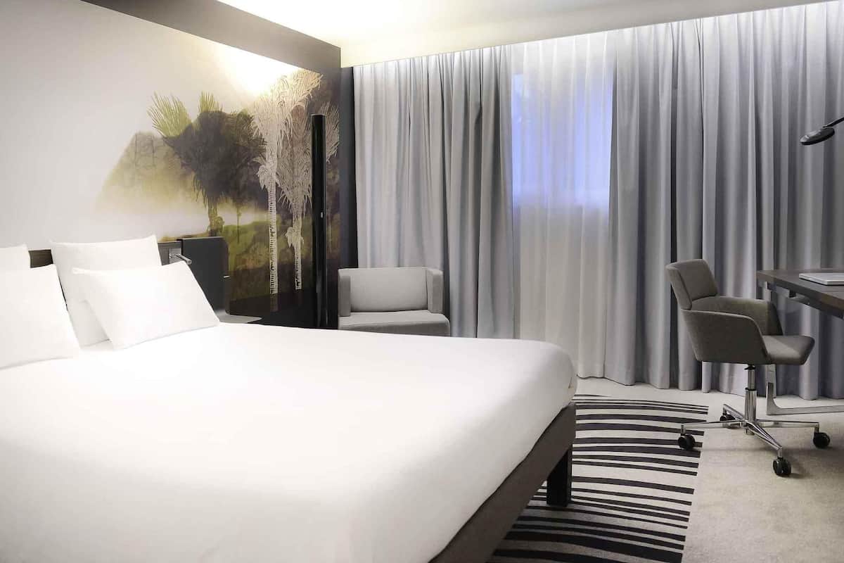 Habitación de hotel con una cama grande, dos sillones, un escritorio y vistas al exterior a través de cortinas translúcidas.