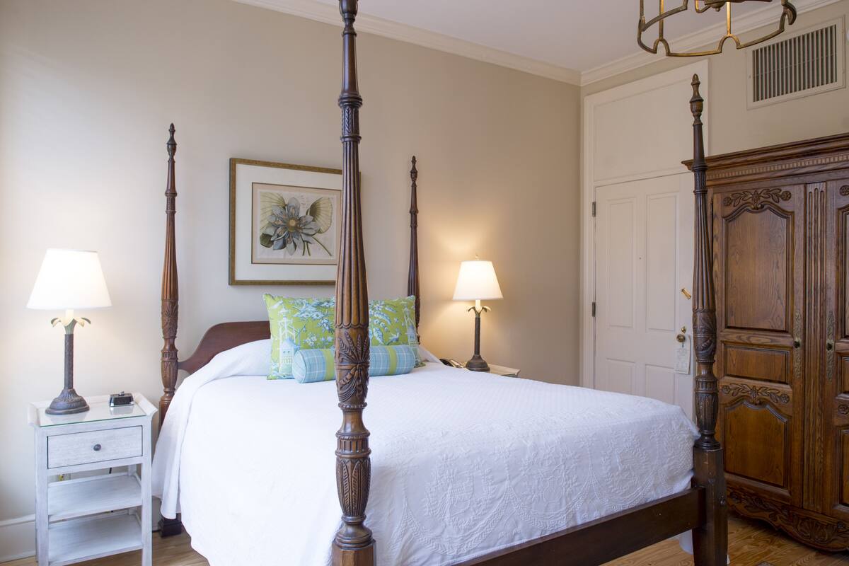 A four-poster bed with a white bedspread, two bedside tables with lamps, and a wooden wardrobe.