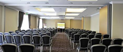 Un salón de conferencias con filas de sillas, suelo alfombrado y una pantalla para proyector.