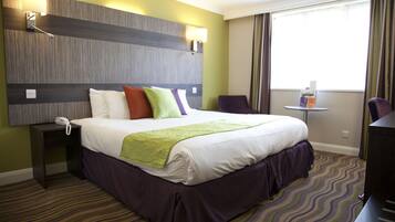 A hotel room with a large bed, a bedside table with a phone, a small table with a magazine, and a chair.