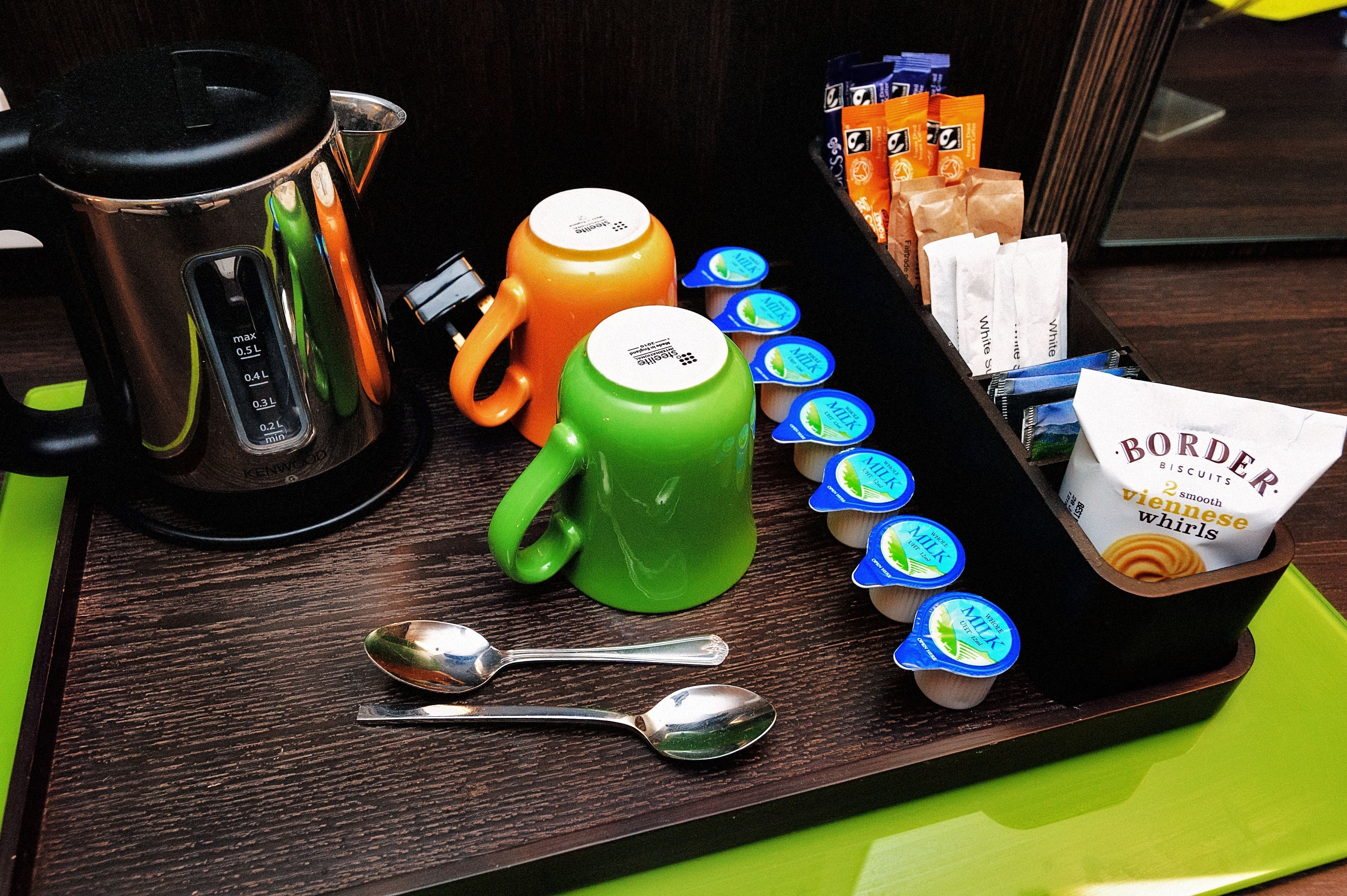 A tray with a coffee maker, two mugs, a set of spoons, and packaged food items.