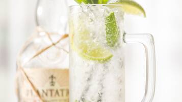 Un grand verre transparent contenant des glaçons et des rondelles de citron vert, des feuilles de menthe et une paille, avec une bouteille portant l’étiquette « PLANTATI » en arrière-plan.