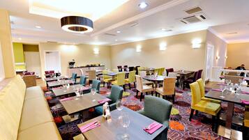 A hotel dining area with multiple tables and chairs, a sofa, and a patterned carpet.