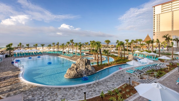 Een zwembadgedeelte van een resort met meerdere zwembaden, palmbomen en een groot gebouw op de achtergrond.