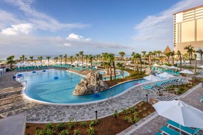 Een zwembadgedeelte van een resort met meerdere zwembaden, palmbomen en een groot gebouw op de achtergrond.