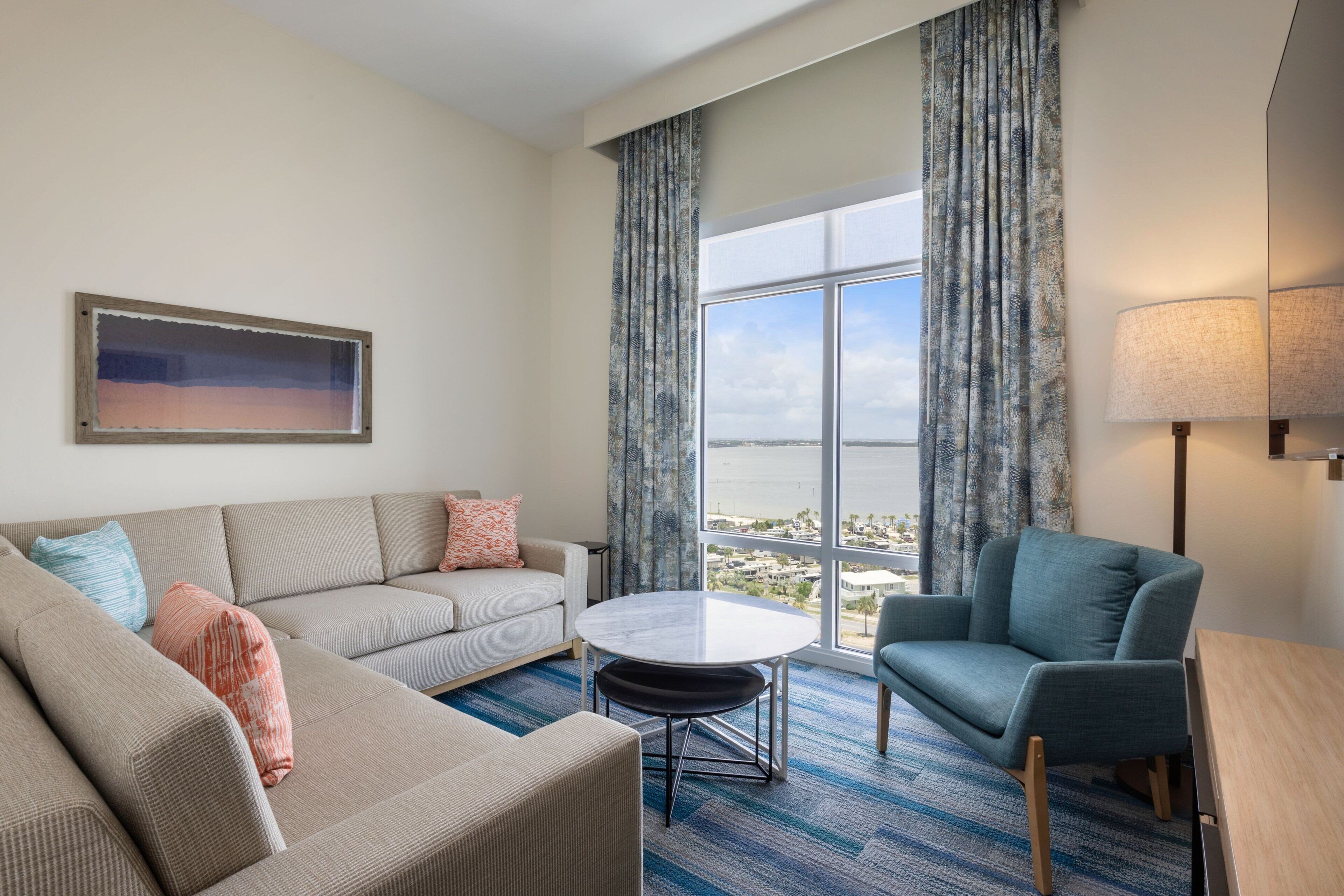 A modern hotel room with a large window, a sofa, a chair, a small table, and a lamp.
