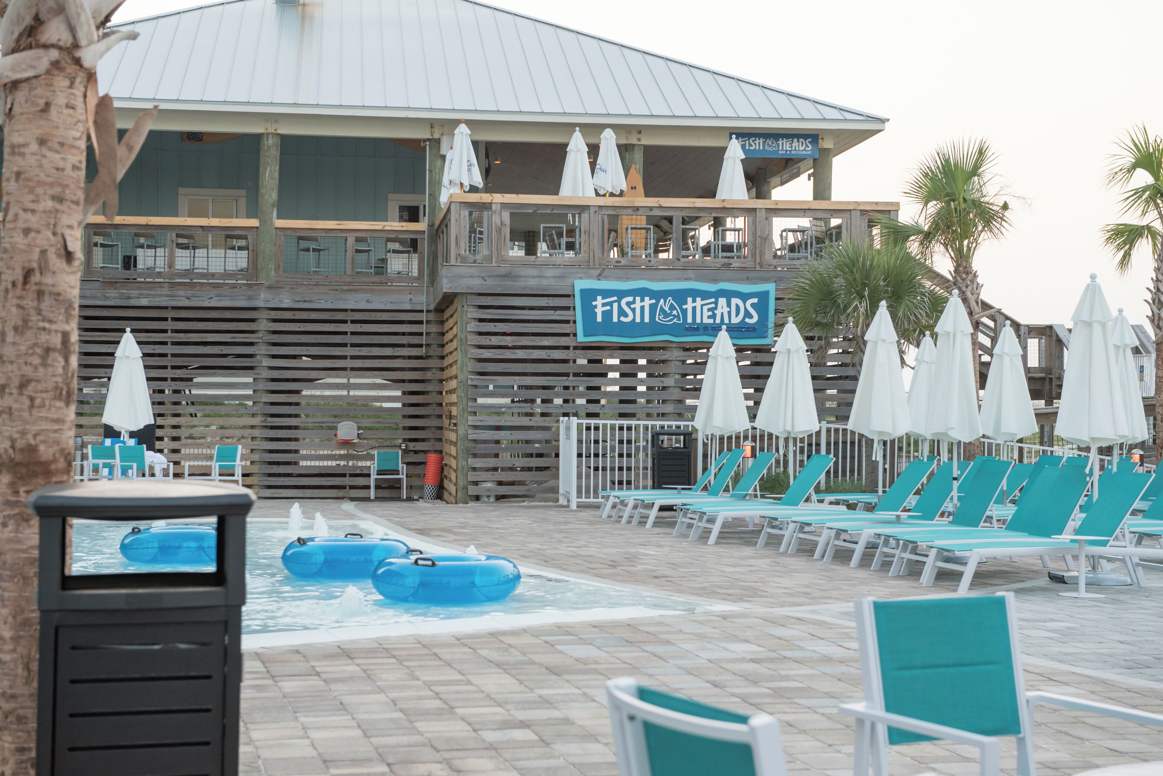 A pool area with lounge chairs and umbrellas, a sign for 'Fist Heads', and a building with a wooden deck.
