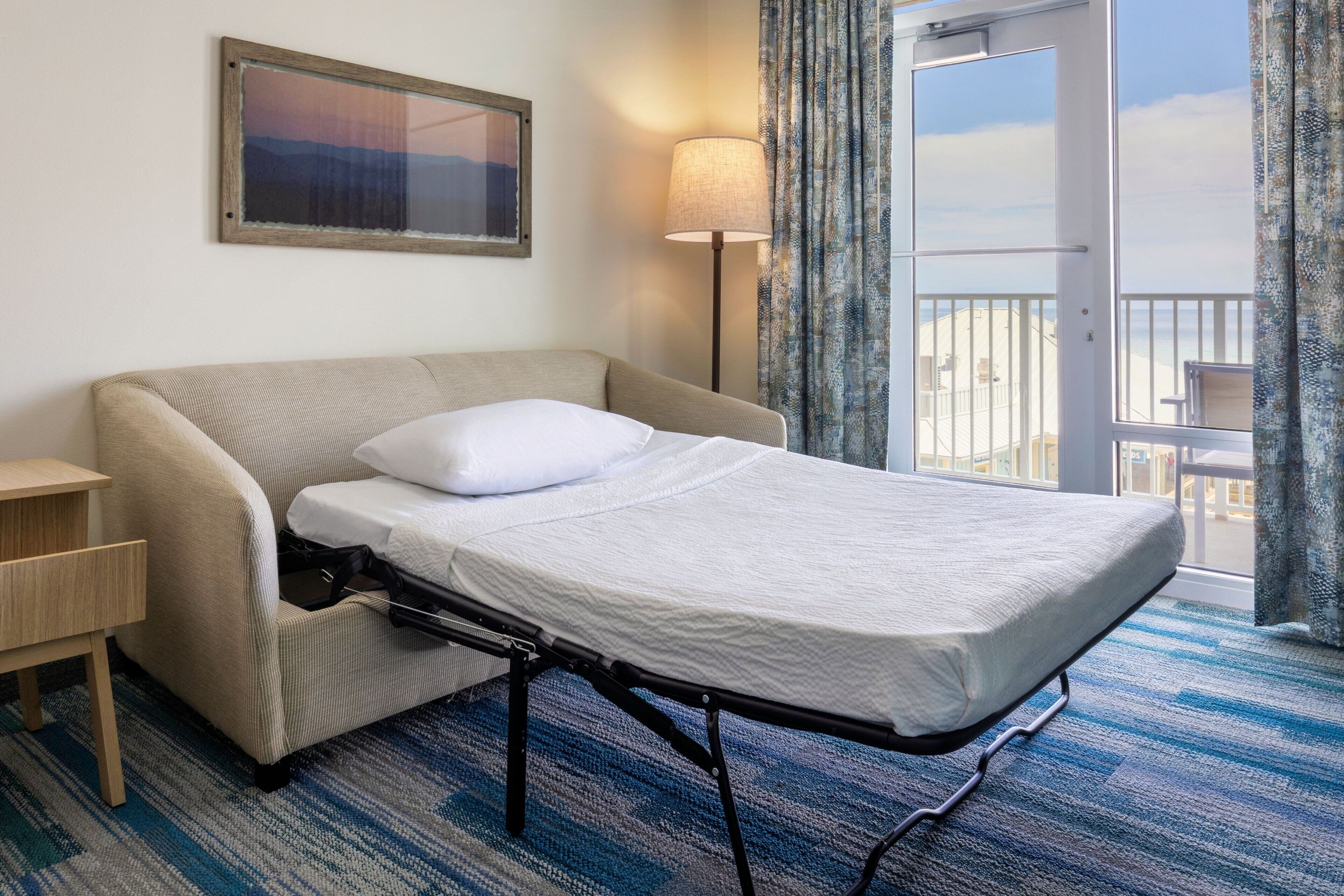 Een kleine hotelkamer met een enkel bed, nachtkastje, lamp en uitzicht op het buitenbalkon.
