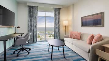 A hotel room with a desk, office chair, sofa, and a view of the city through large windows.