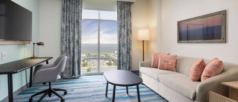 A hotel room with a desk, office chair, sofa, and a view of the city through large windows.