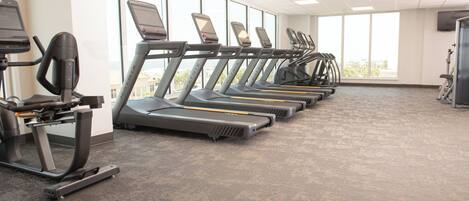 A gym with treadmills aligned in a row, large windows, and a carpeted floor.