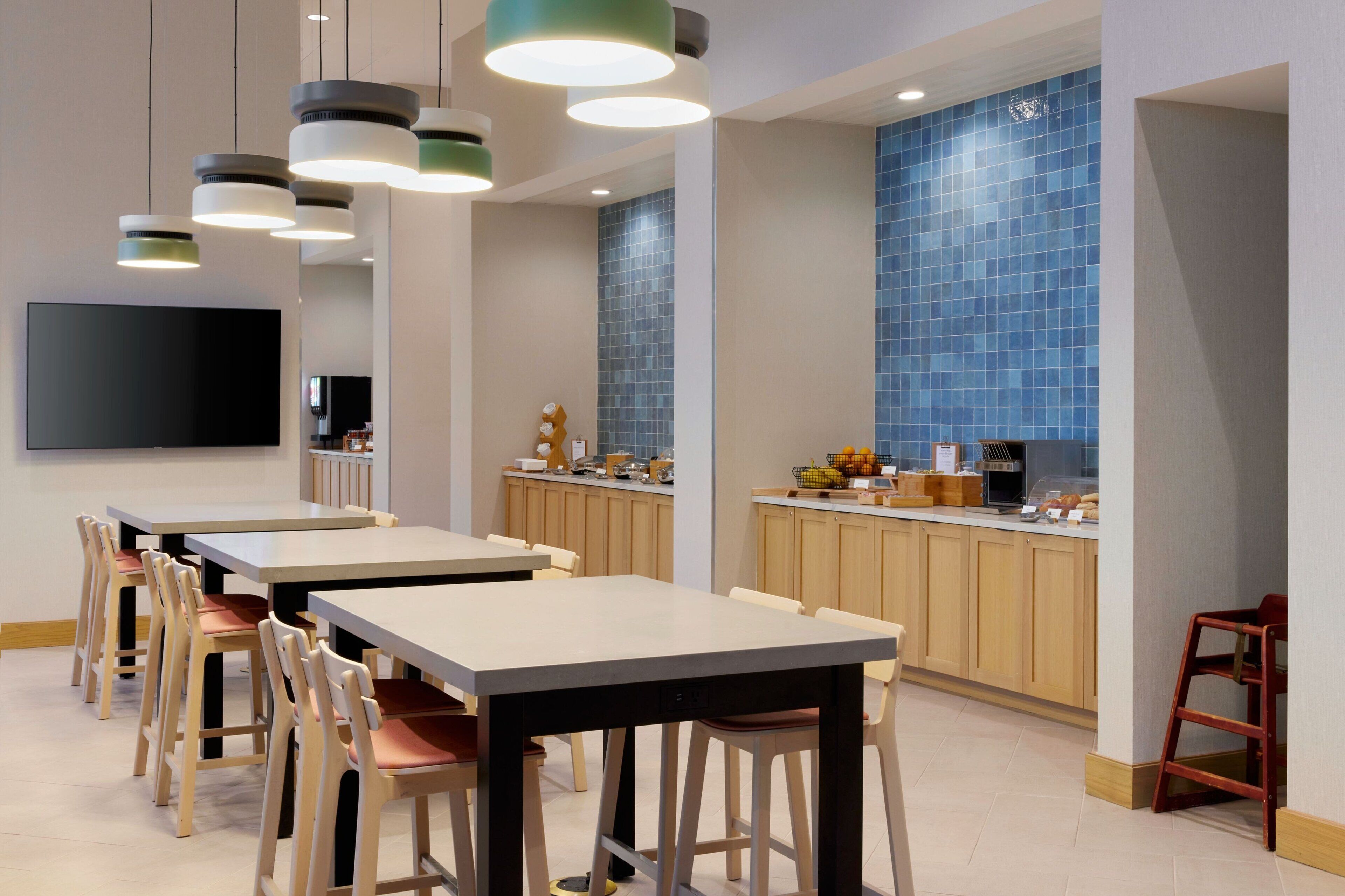 Un comedor moderno con varias mesas y sillas, una encimera de cocina con diversos objetos, y una gran pared de azulejos azules.