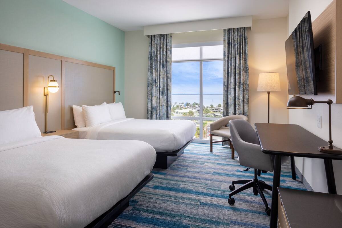 A hotel room with two beds, a desk, a chair, and a large window with curtains.