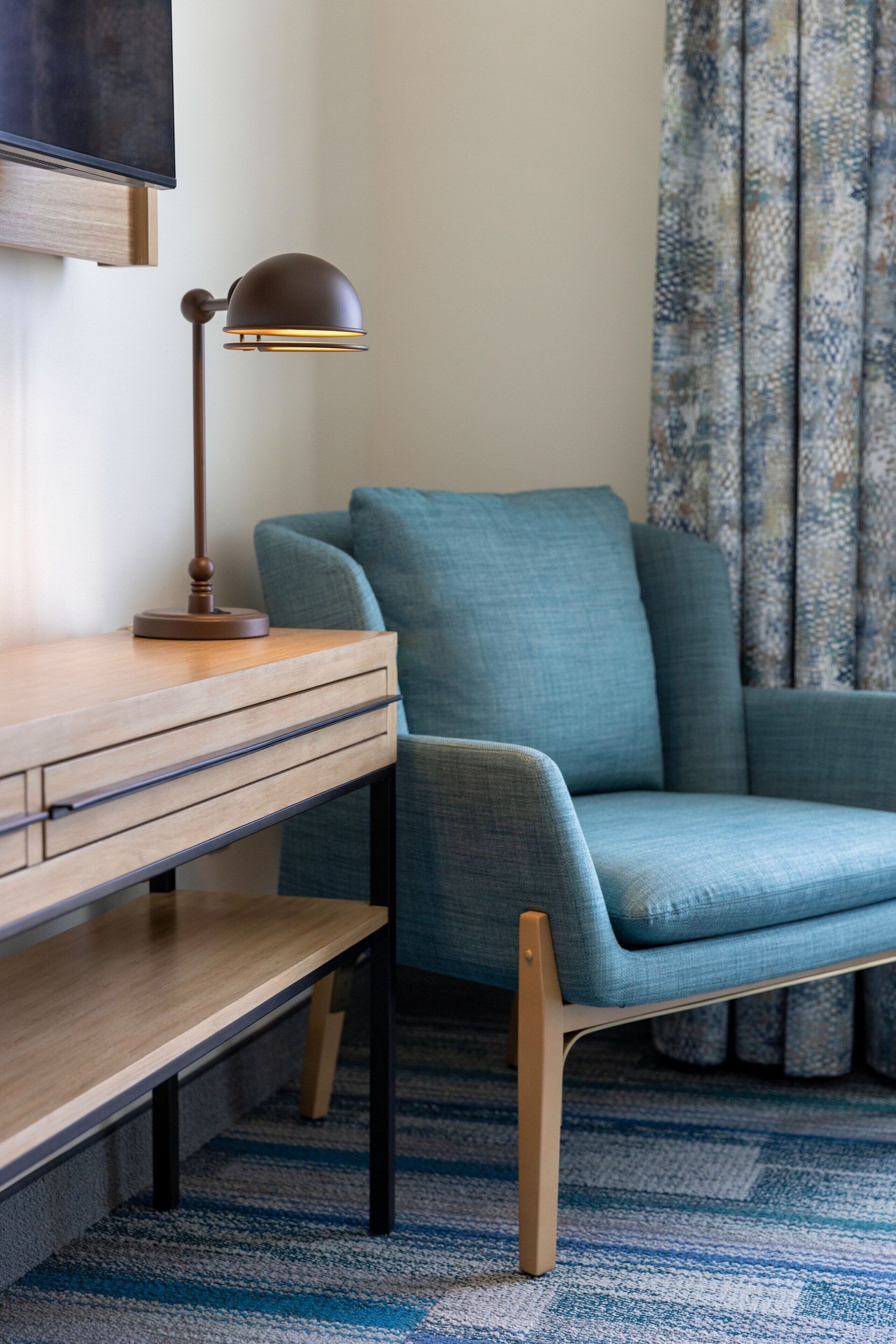 A room with a wooden desk, a blue armchair, a floor lamp, and a patterned curtain.
