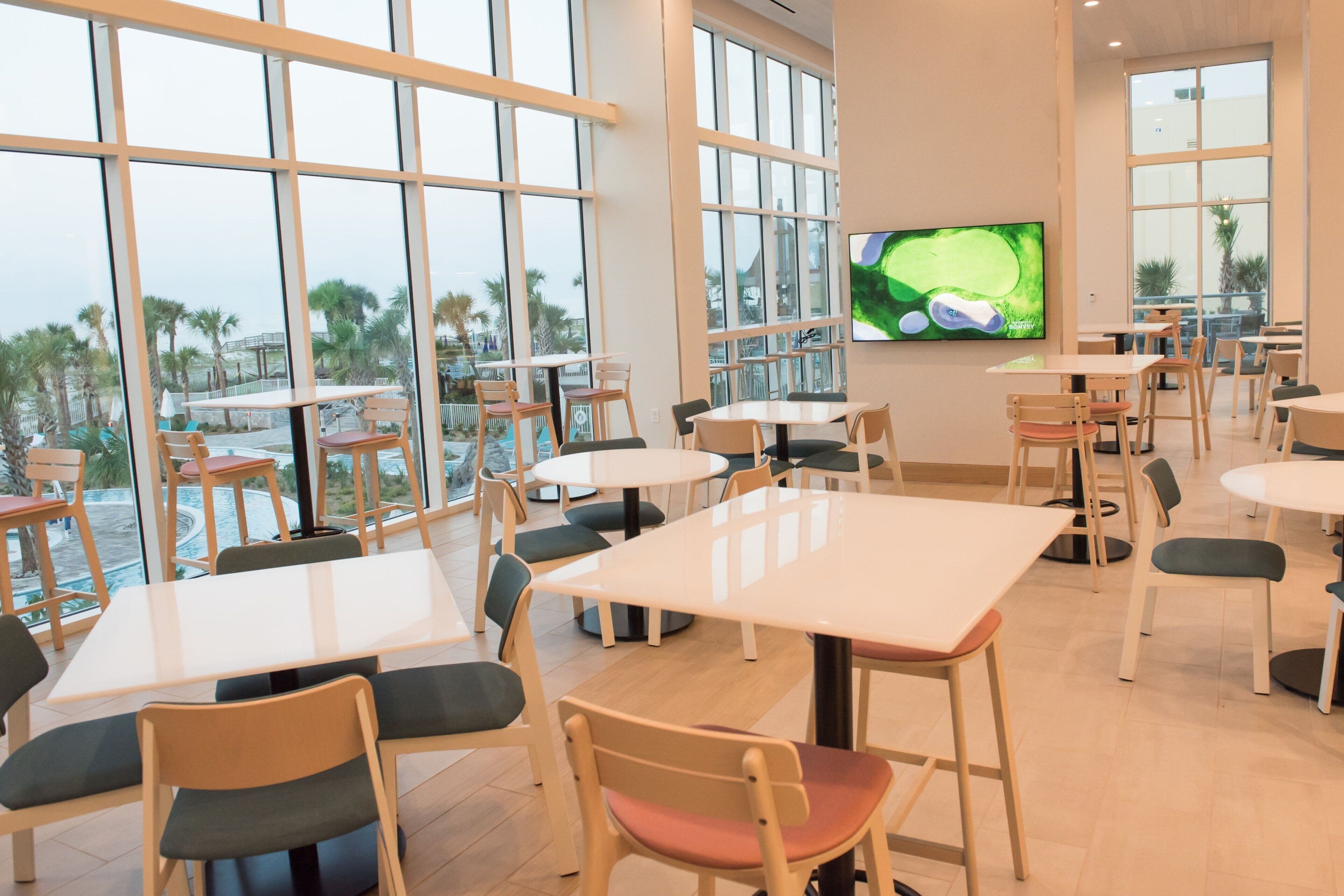 Un espace intérieur spacieux, doté de nombreuses tables et chaises, de grandes fenêtres offrant une vue sur les palmiers et une piscine, ainsi que d’un téléviseur à écran plat fixé au mur.