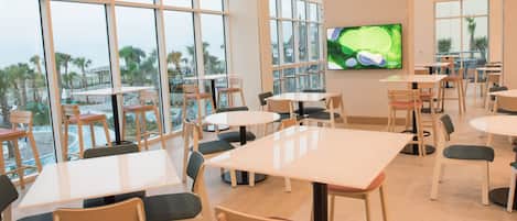 Un amplio espacio interior con varias mesas y sillas, ventanas grandes que ofrecen vistas a palmeras y una piscina, y un televisor de pantalla plana montado en la pared.