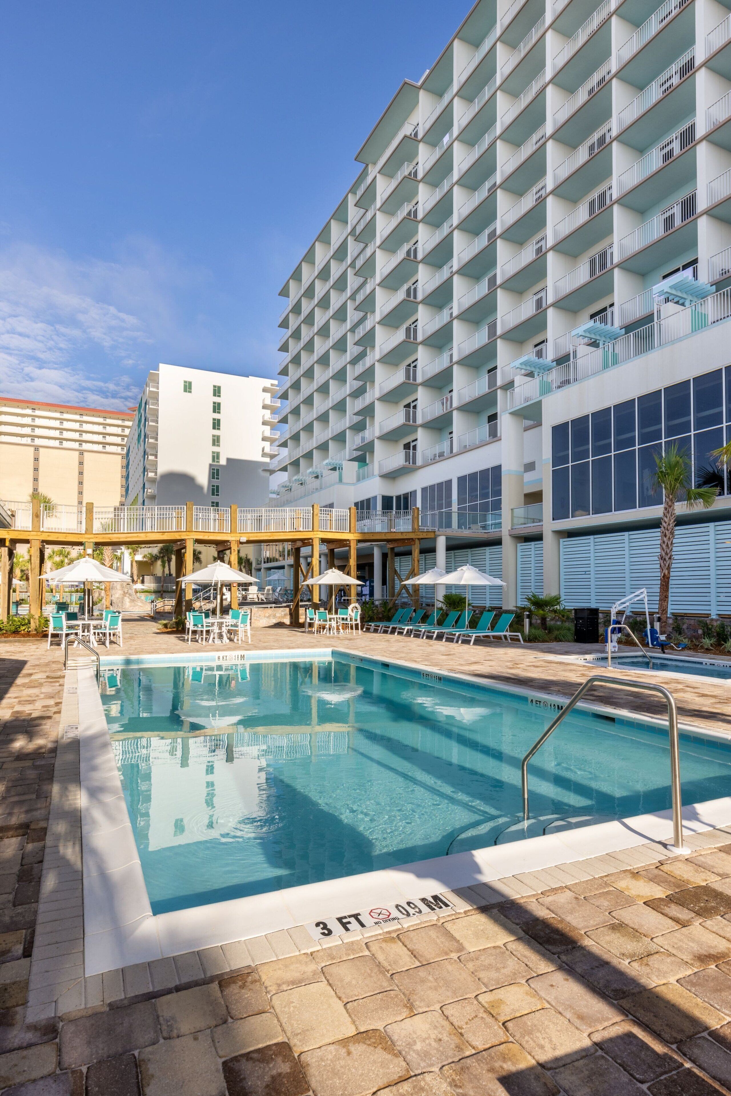 A hotel pool area with lounge chairs and umbrellas, a 3 FT depth sign, and a large building with balconies.