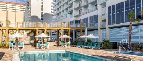 A hotel pool area with lounge chairs and umbrellas, a 3 FT depth sign, and a large building with balconies.