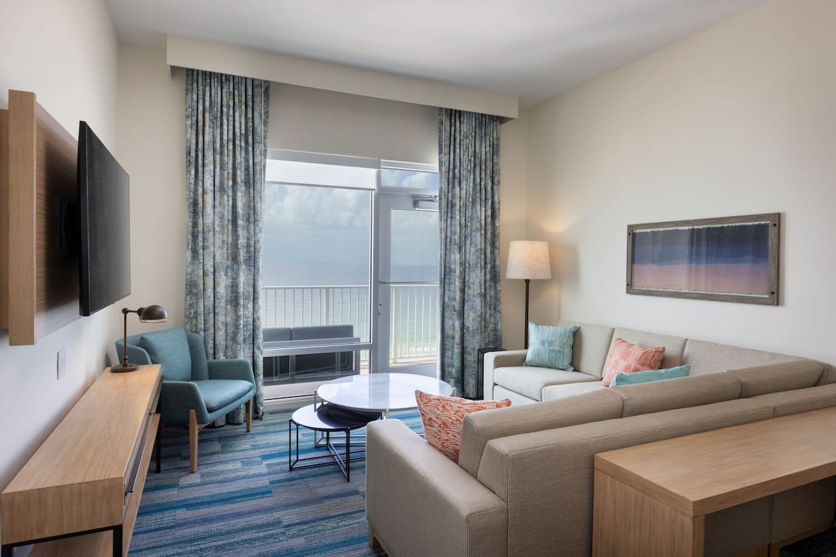 Ein modernes Hotelzimmer mit einer Couch, einem kleinen Tisch, einem Fernseher und einem Balkon mit Vorhängen.