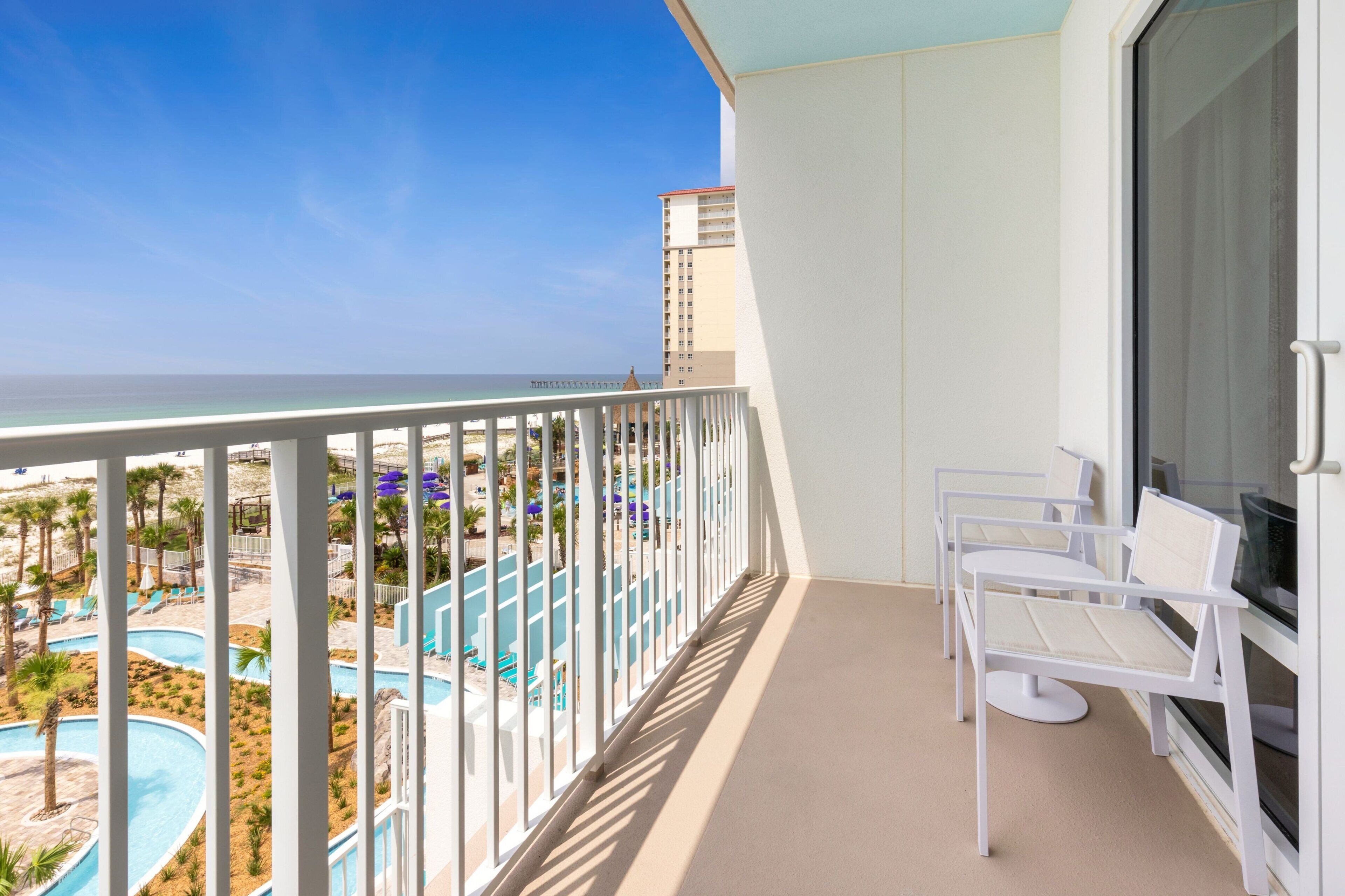 A balcony with a view of a pool and beach, white chairs, and a large window.