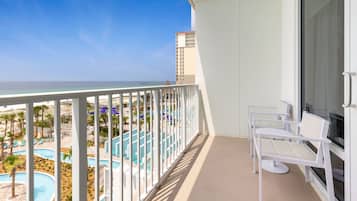 A balcony with a view of a pool and beach, white chairs, and a large window.