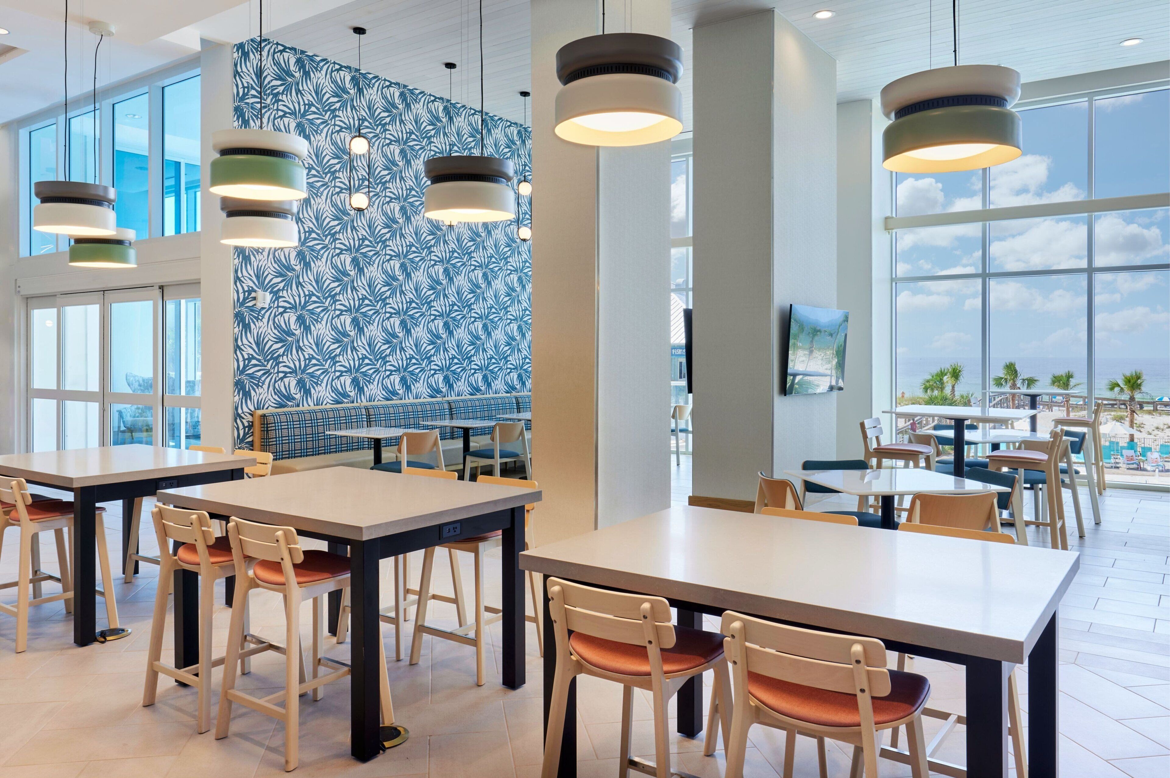 A modern dining area with high ceilings, large windows, and a patterned wall. Tables and chairs are arranged for dining.