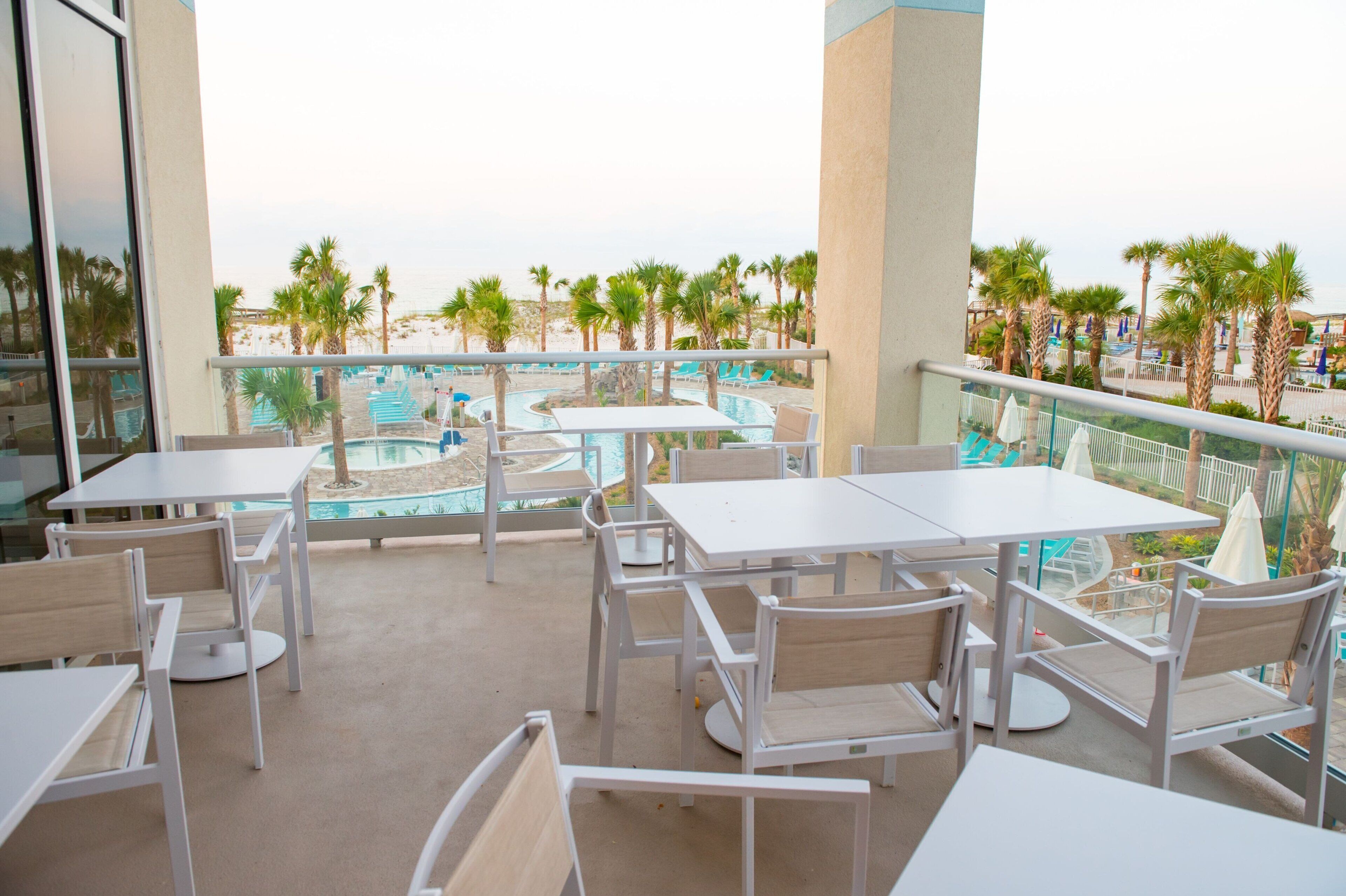 A balcony with white tables and chairs, overlooking a pool area with palm trees and lounge chairs.