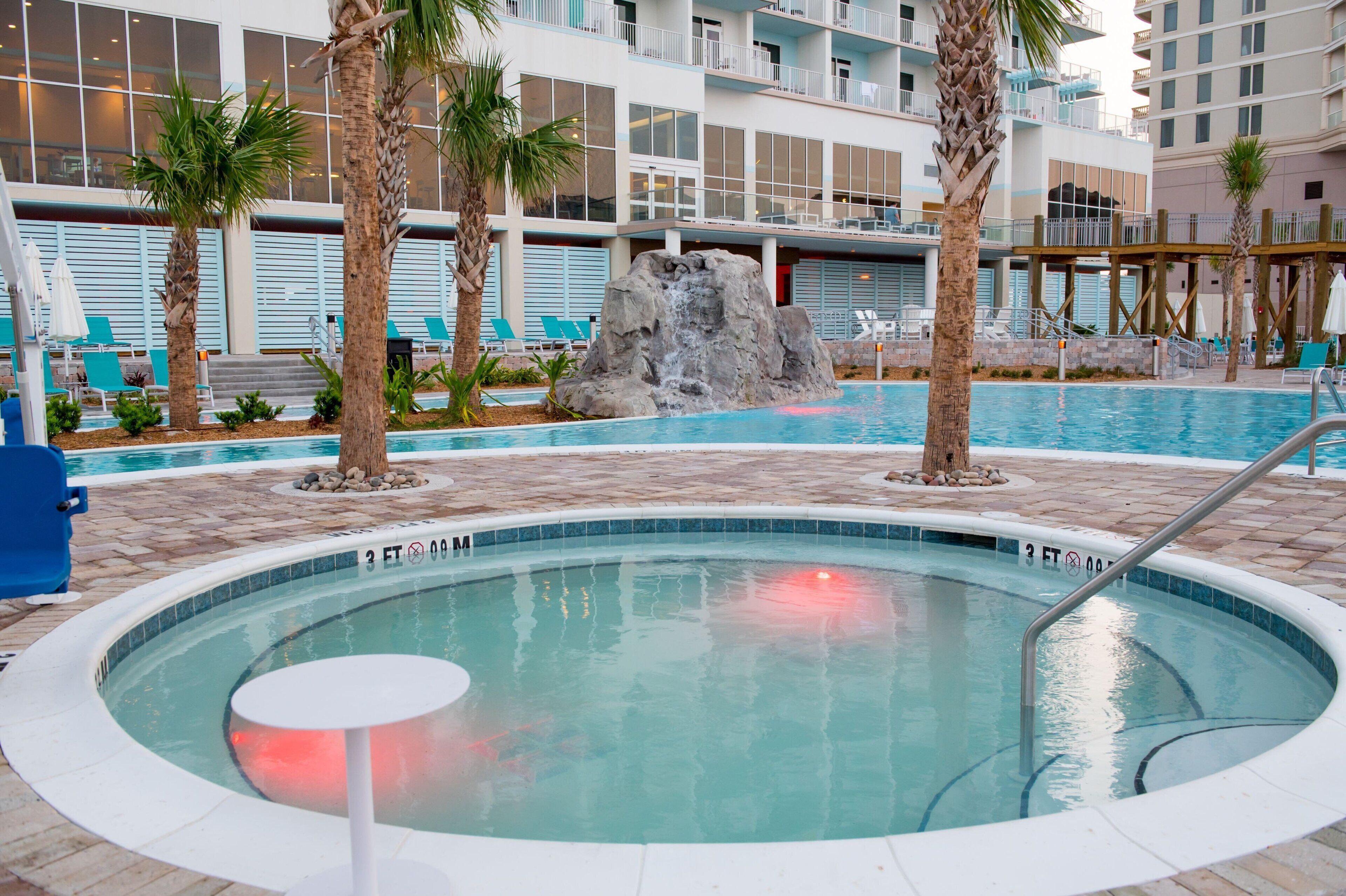 A hotel pool area with a hot tub, palm trees, lounge chairs, and a rocky waterfall feature.