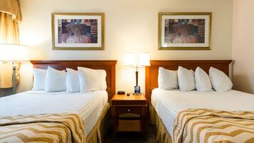 A hotel room with two beds, each with white bedding and striped blankets. There are two bedside tables with lamps, a nightstand, and two framed pictures on the wall.