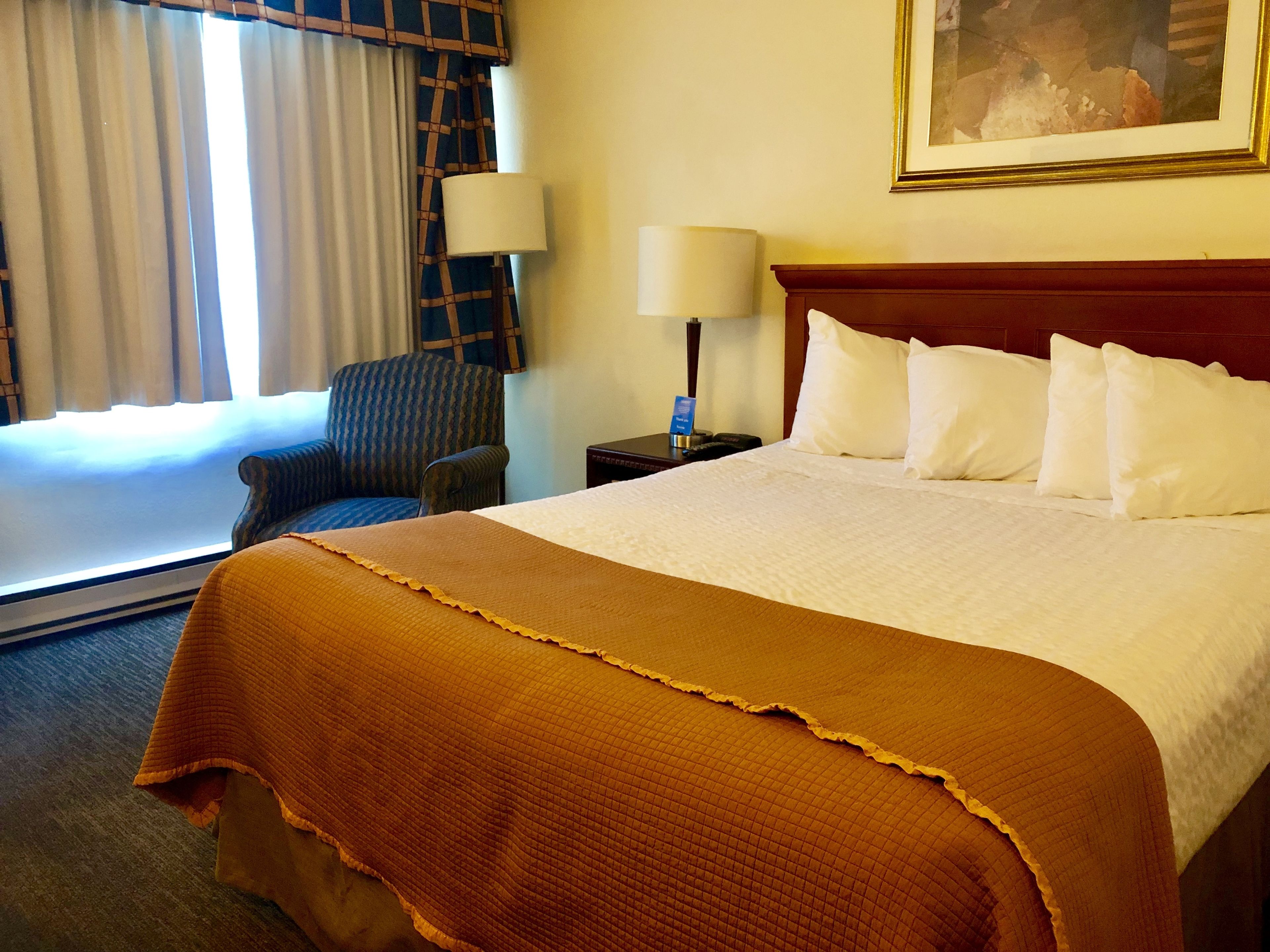 A hotel room with a large bed, a chair, a nightstand with a lamp, and a window with curtains.