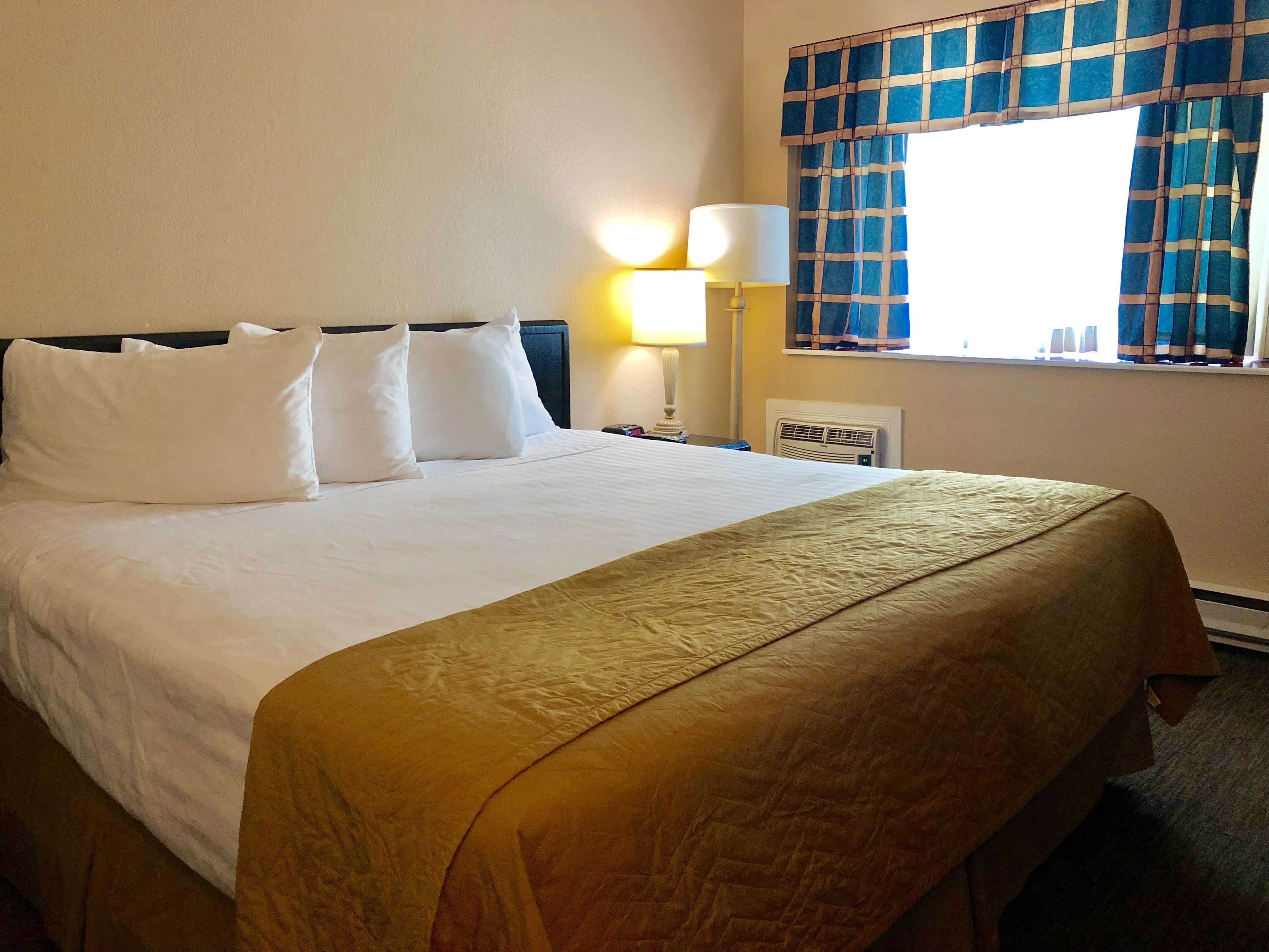 Habitación de hotel con una cama grande, ropa de cama blanca, cabecera, lámpara de noche, ventana con cortinas azules y un calefactor de pared.