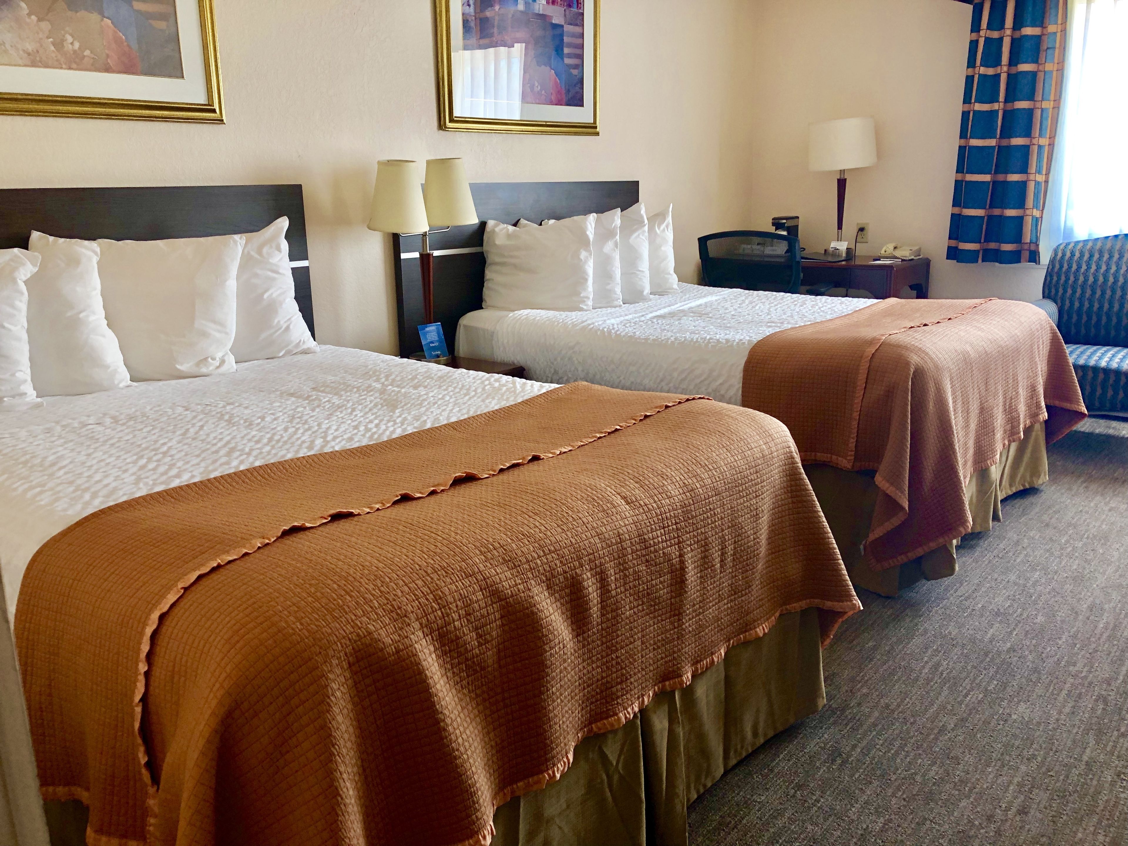 A hotel room with two beds, each with white linens and a brown blanket. There are two bedside tables with lamps, a chair, and a blue curtain.