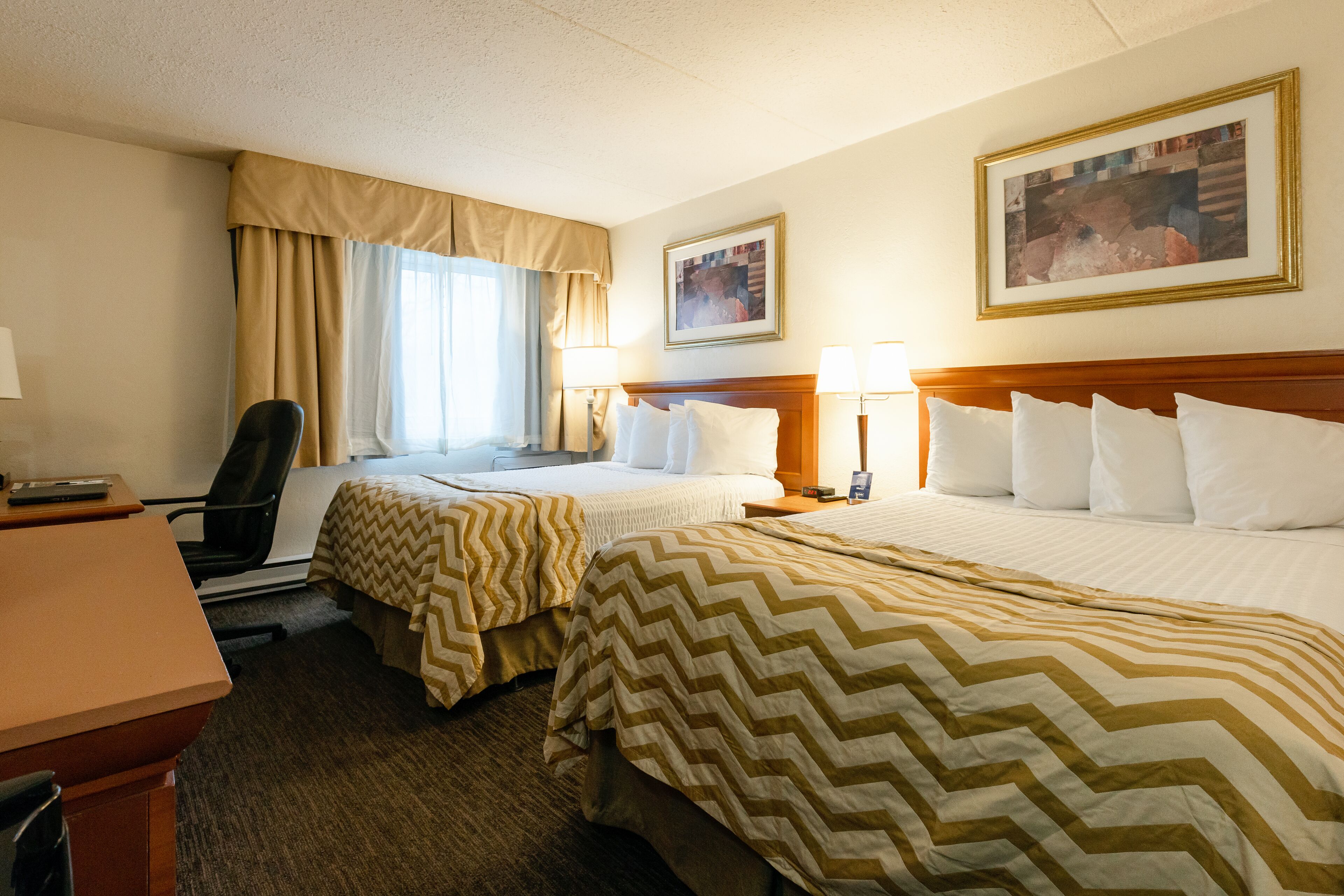 A hotel room with two beds, a desk, and a chair. There are two lamps on the nightstands and a framed picture on the wall.
