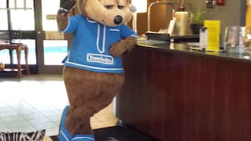 Un personaje publicitario con uniforme azul de pie en un mostrador de recepción con un tapete de 'Travelodge' delante.