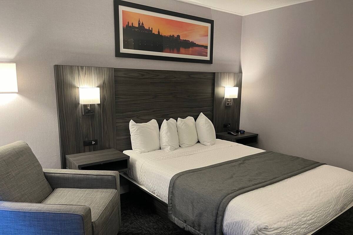 Une chambre d’hôtel avec un lit, deux lampes de chevet, une chaise, une table de chevet et un tableau encadré au mur.