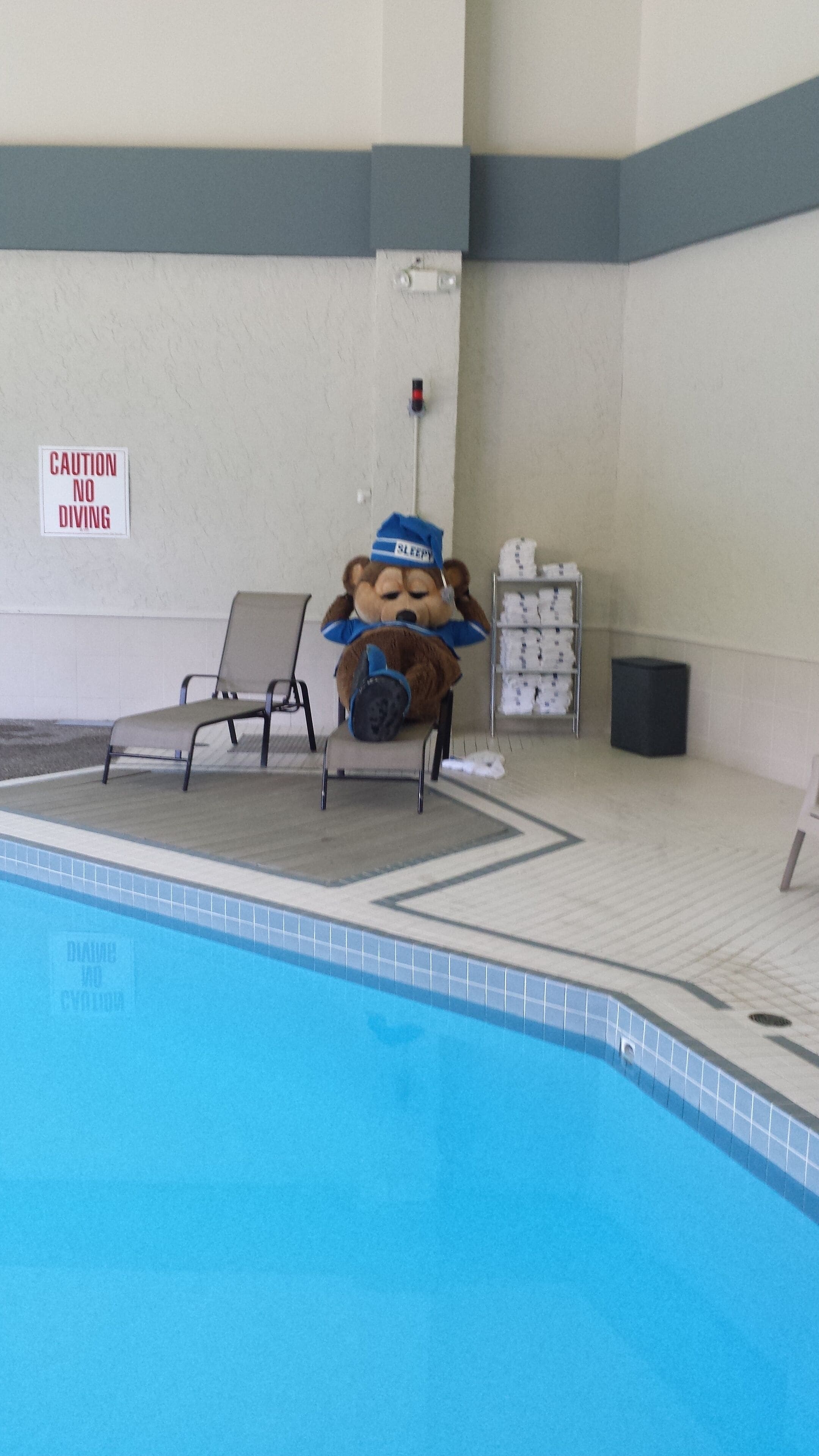 A pool area with a caution sign, a lifeguard mascot, lounge chairs, and a tiled floor.