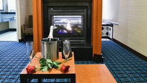 A room with a fireplace, a wooden table with a bottle of champagne, two glasses, and a bouquet of roses. - Fairfield Inn & Suites Hartford Airport (Windsor Locks)