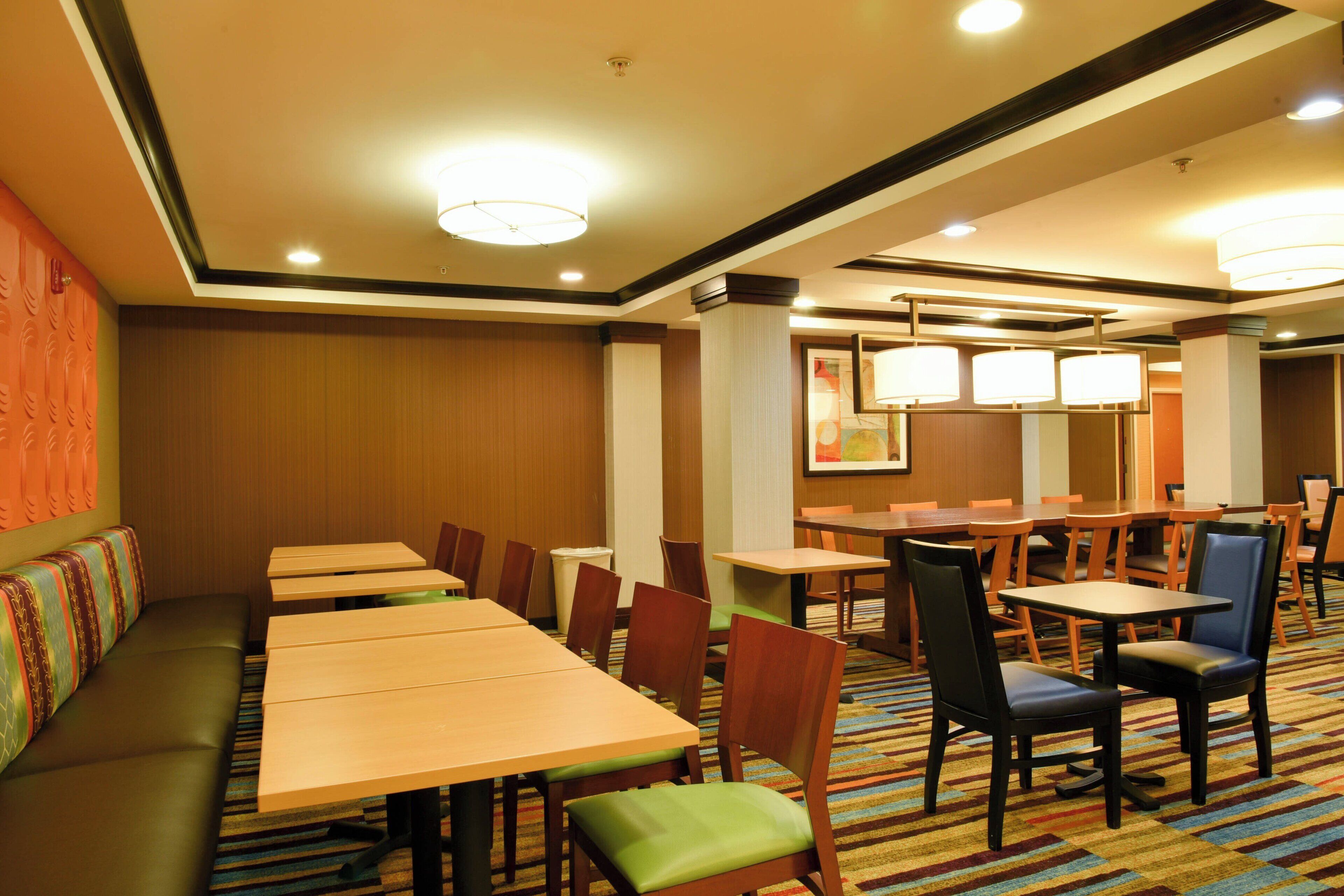 A hotel dining area with multiple tables and chairs, a long bench, and a carpeted floor.