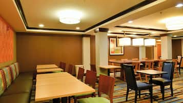 A hotel dining area with multiple tables and chairs, a long bench, and a carpeted floor.