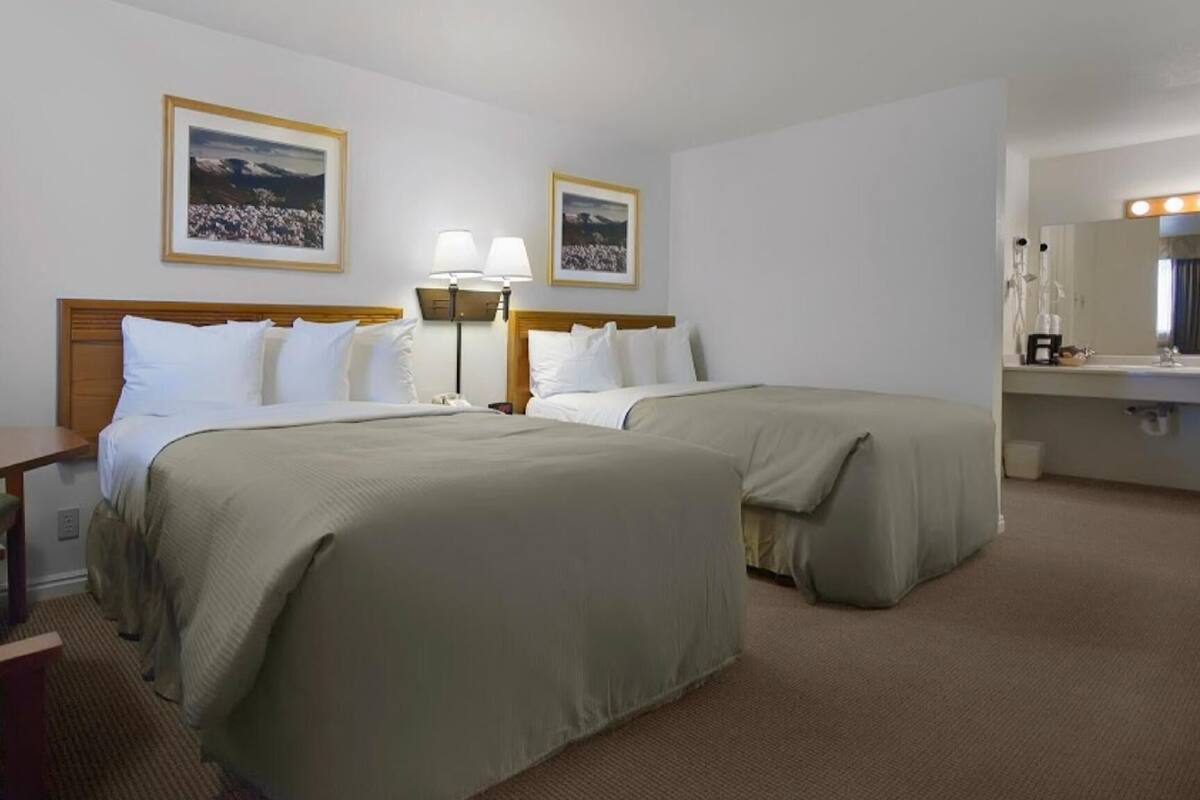 Quarto de hotel com duas camas, uma escrivaninha, uma cadeira, abajur, espelho e quadros na parede.