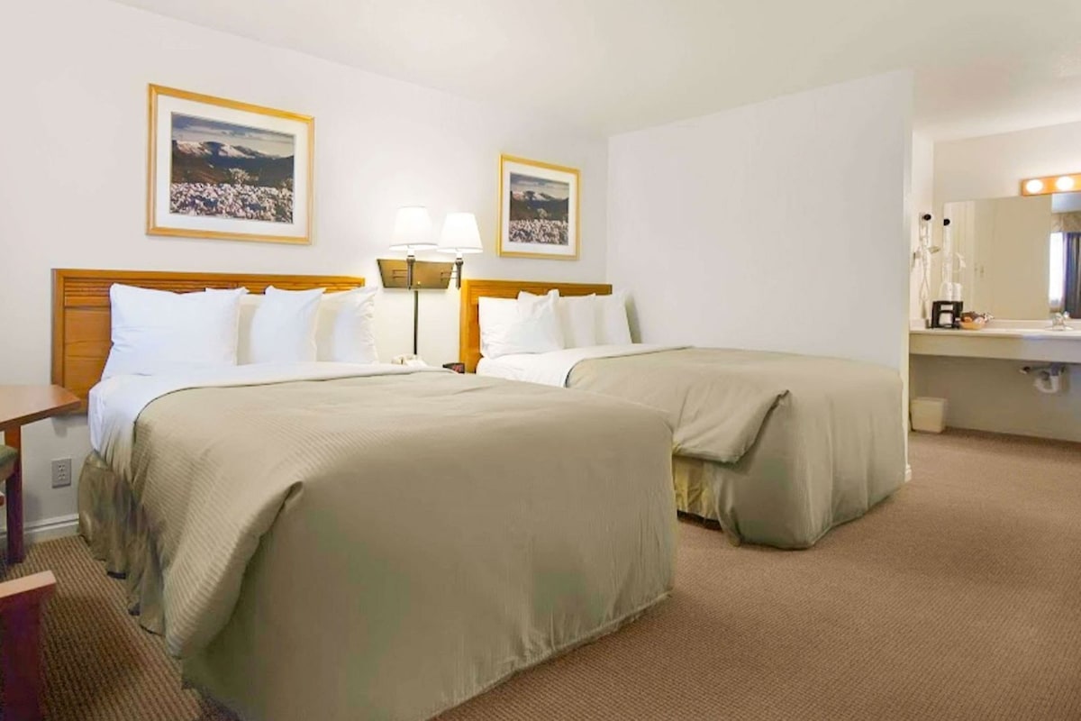 A hotel room with two beds, a desk, a chair, a lamp, a mirror, and two framed pictures on the wall.