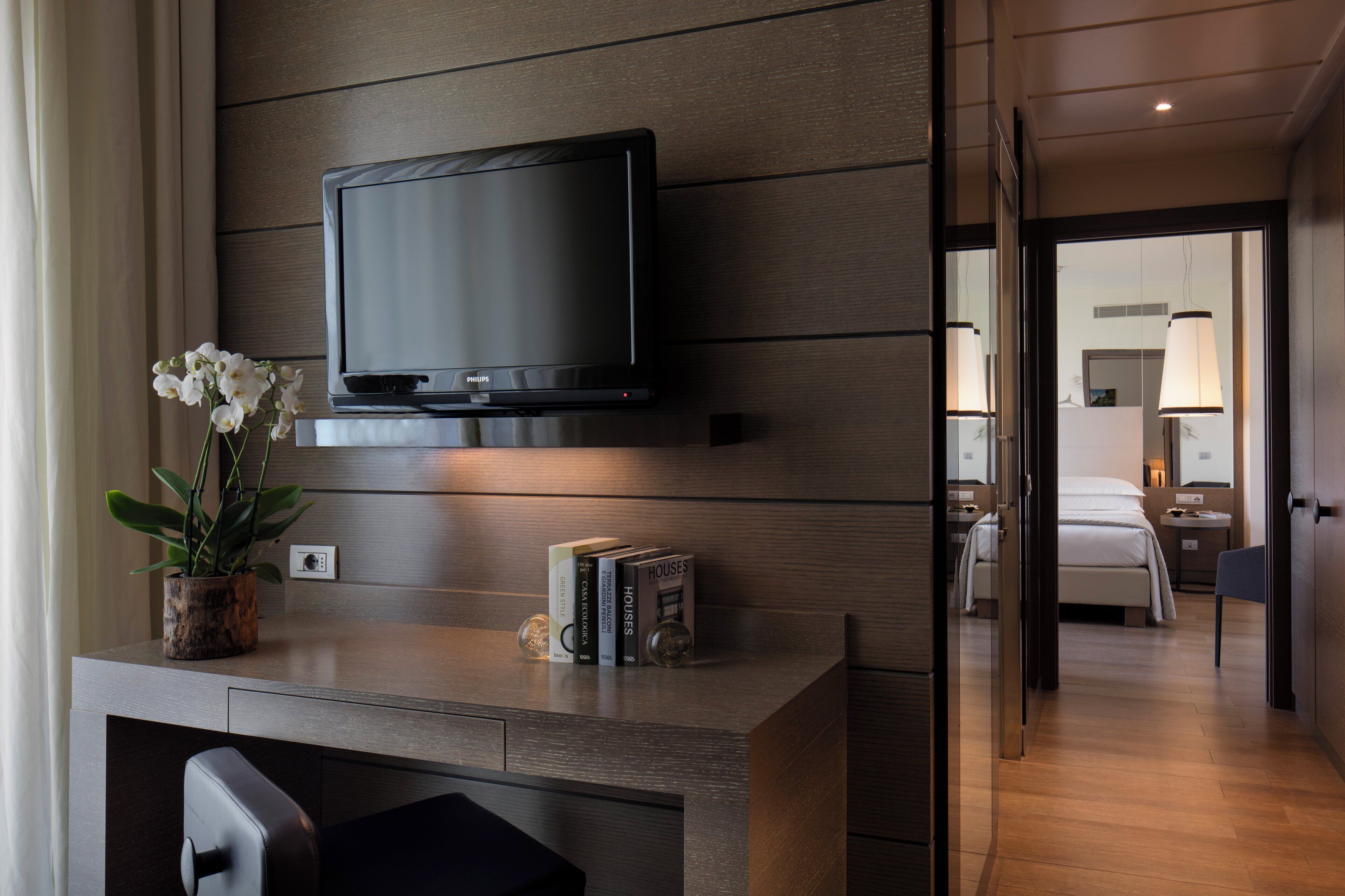 A hotel room with a flat-screen TV mounted on the wall, a small table with a potted plant, and a bedside table with books.